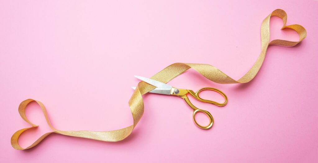 Gold scissors cutting golden ribbon ending in two hearts, pink background, top view.
